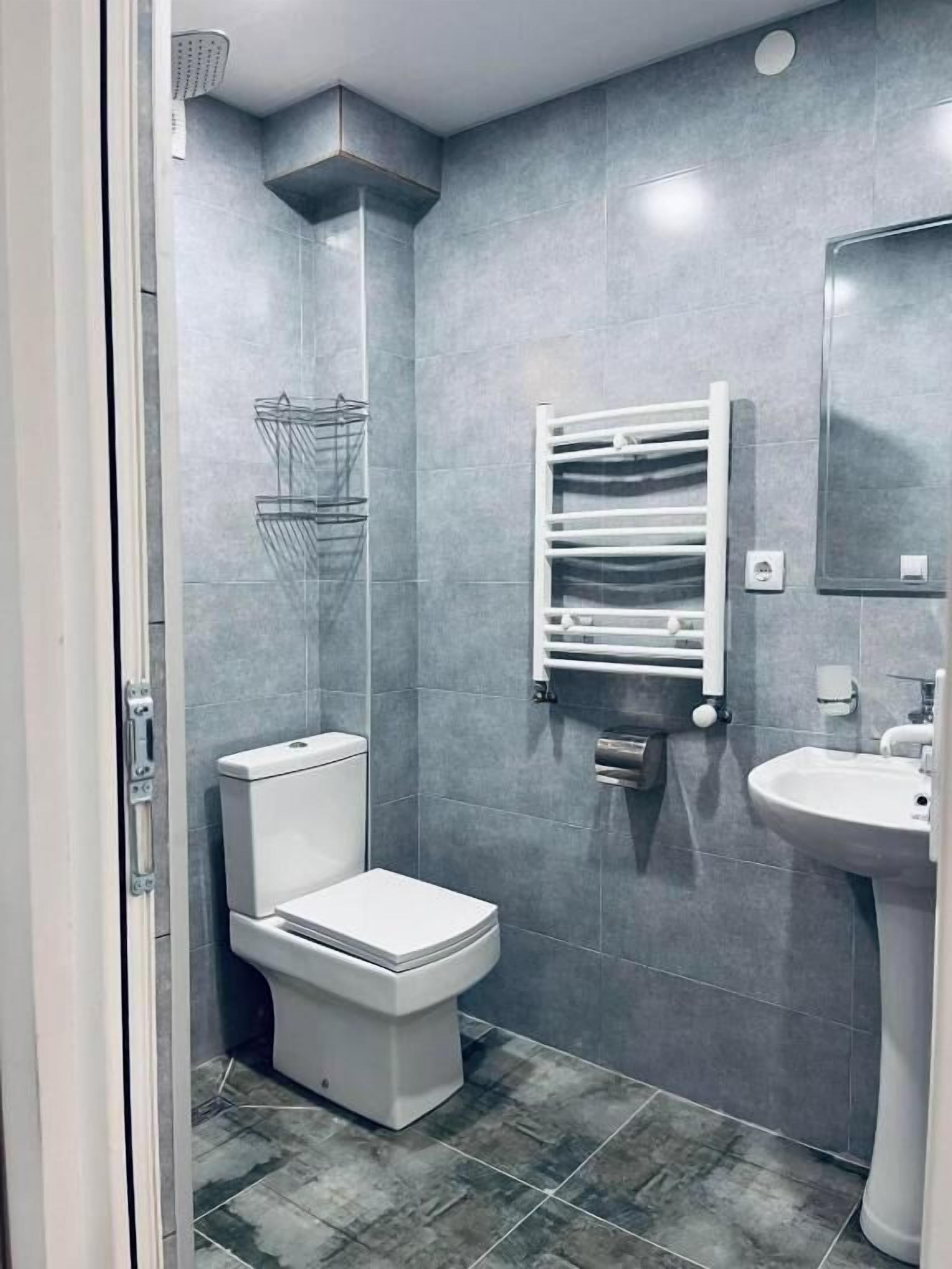 Un baño con azulejos grises, un inodoro, un lavamanos, un perchero para toallas y una ducha.