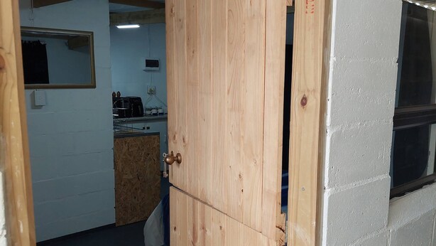 A room with a wooden door, a window, and a kitchen area in the background.