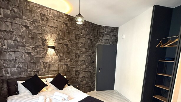 A bedroom with a stone wall, a bed with a black headboard, a bedside table, a hanging light fixture, a wardrobe with a mirror, and a door. - bütünoğlu pansiyon (Antalya)