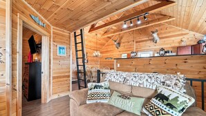 Interior de una cabaña de madera con sofá, almohadones, una lámpara de araña y trofeos de animales colgados.