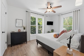 Une chambre à coucher avec un lit, un ventilateur de plafond, une télévision et une fenêtre avec des rideaux.