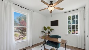 Ein Zimmer mit Schreibtisch aus Holz, einem Türkis-farbenen Stuhl, einem Deckenventilator und großen Fenstern mit weißen Vorhängen.