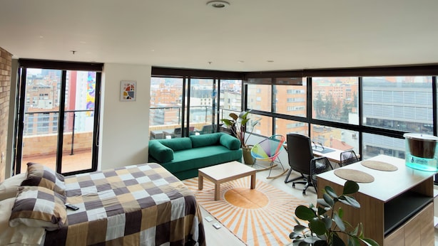 Habitación de hotel moderna con una cama grande, un sofá verde, un escritorio y vistas a la ciudad a través de amplias ventanas.