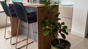 Una cocina moderna con una isla de madera, dos sillas negras, una planta en maceta y vistas de edificios a través de amplios ventanales.