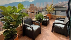 Un balcón con mobiliario de mimbre, plantas en macetas y vistas a la ciudad.