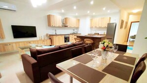 Une cuisine moderne avec des meubles en bois, un coin repas avec une table dressée pour deux personnes, et un téléviseur à écran plat fixé au mur.