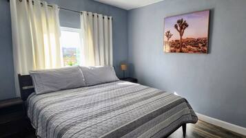 A bedroom with a bed, curtains, a ceiling fan, a nightstand, a lamp, and a desert landscape painting.