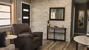 Un salon avec un fauteuil marron, une petite table ronde, une table d’appoint agrémentée d’objets décoratifs, un miroir et un mur dont le motif rappelle celui d’un mur en briques.