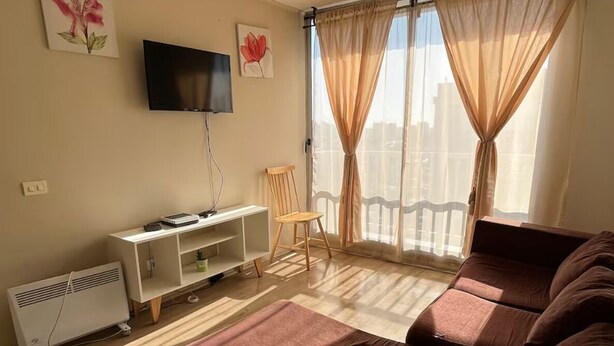 A living room with a sofa, a TV, a radiator, a chair, and a balcony with curtains.