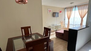 A dining area with a glass-top table and wooden chairs, a sofa, a TV on the wall, and a balcony with curtains.
