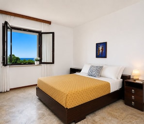Una camera da letto con un letto, un comodino, una lampada, un quadro, una finestra con tende e vista mare.