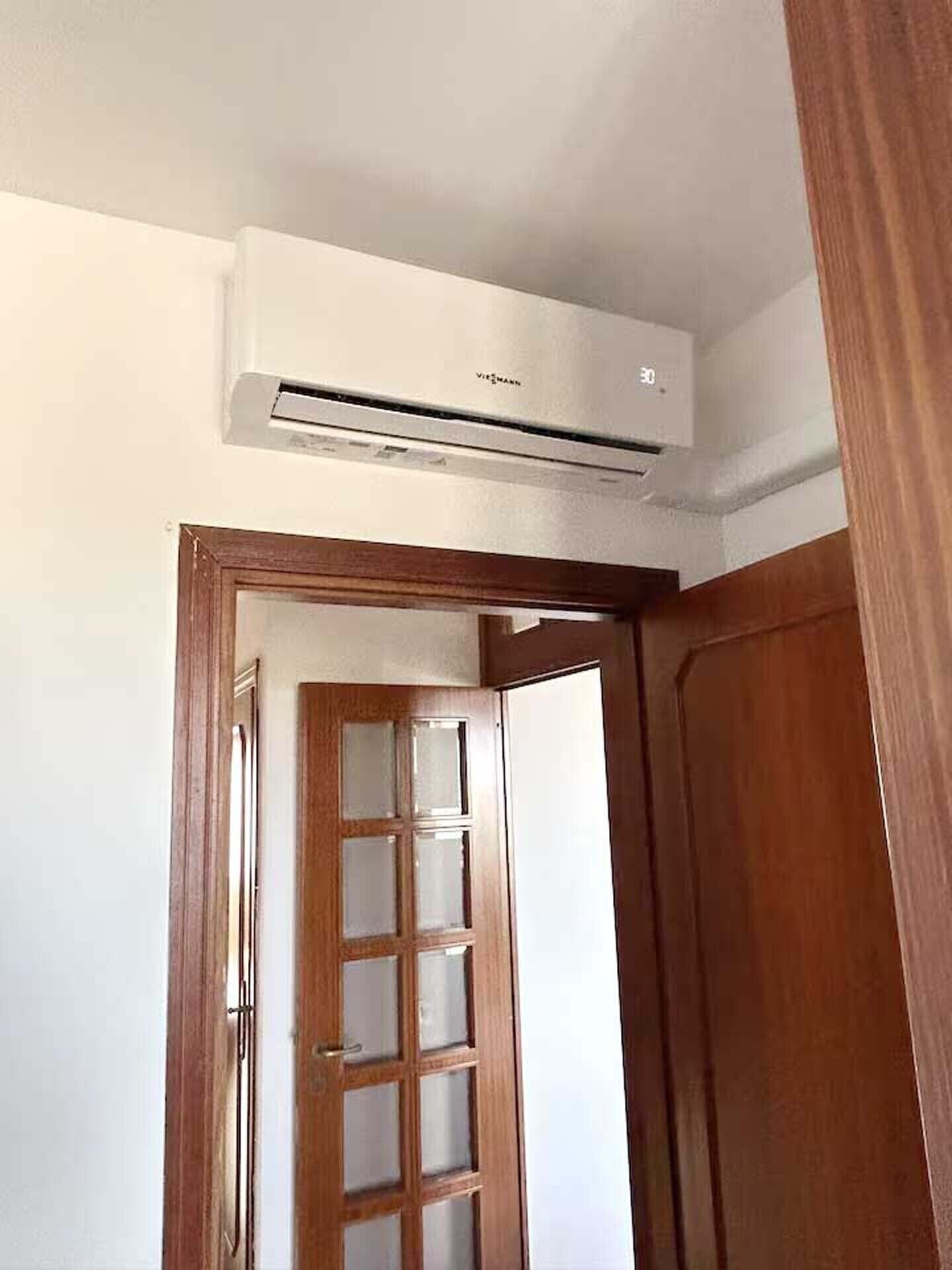 Una habitación con puerta de madera, pared blanca y unidad de aire acondicionado blanca montada encima de la puerta.