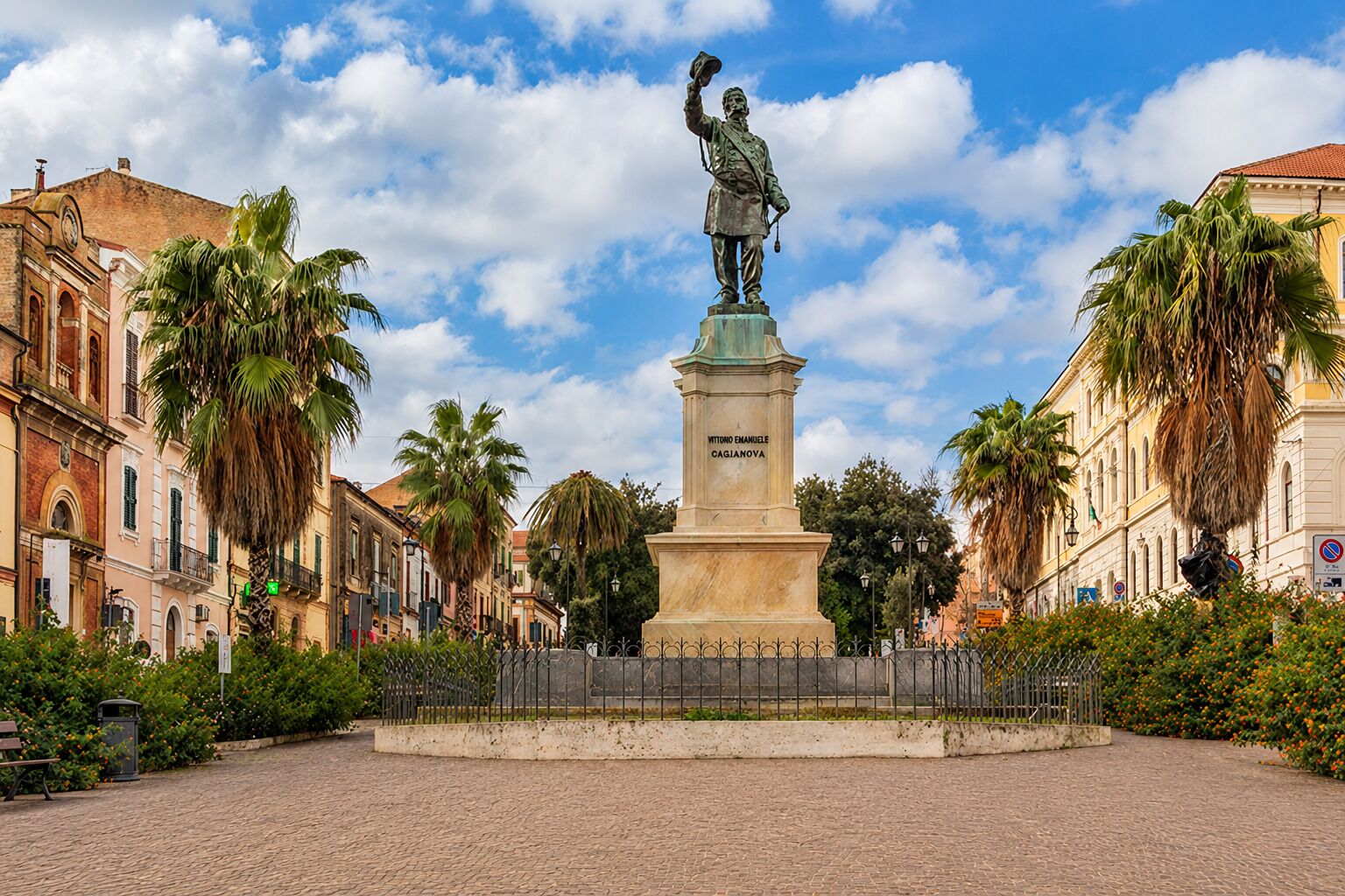 A statue in a square with cobblestone pavement, surrounded by historic buildings and palm trees.