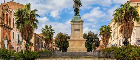 A statue in a square with cobblestone pavement, surrounded by historic buildings and palm trees.