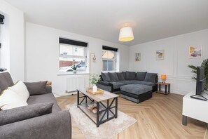 Ein modernes Wohnzimmer mit grauem Sofa, einem Holztisch, einer grauen Eckcouch, einem weißen Schreibtisch und einem Fernseher.