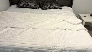 Una cama con colcha blanca, dos almohadas estampadas, un cabecero de madera y una mesita de noche con lámpara.