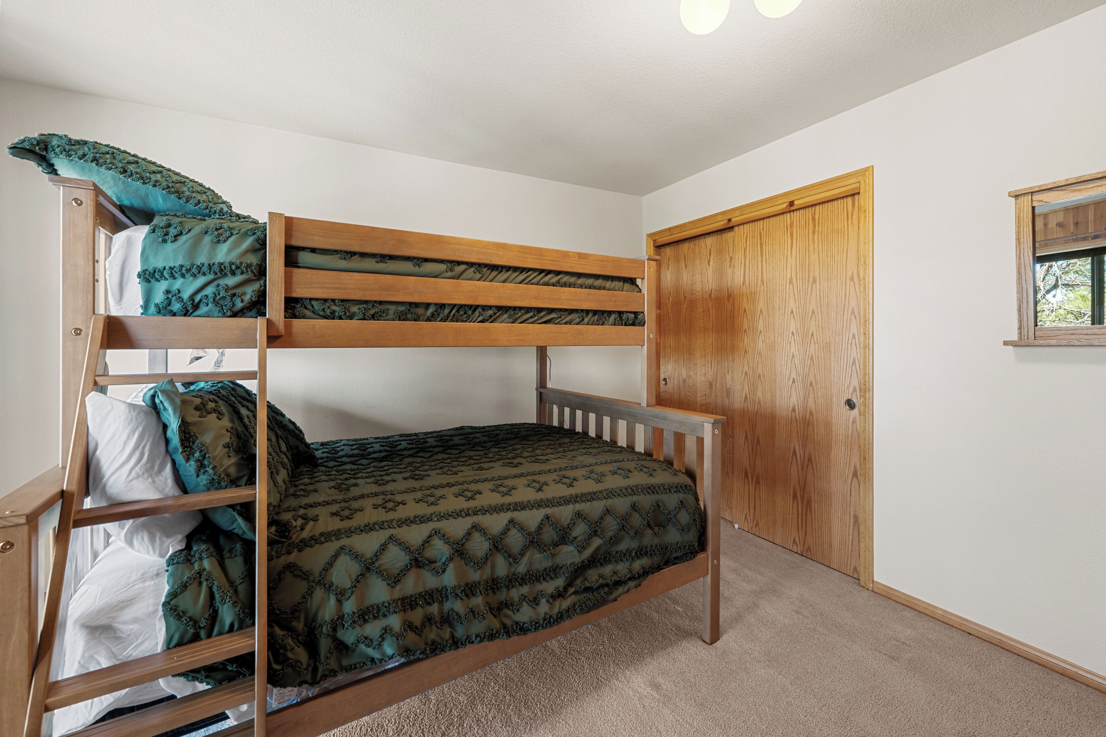 Ein Stockbett mit einem gemusterten Bettbezug, ein Holzschrank und ein Fenster mit Blick auf Grünflächen.