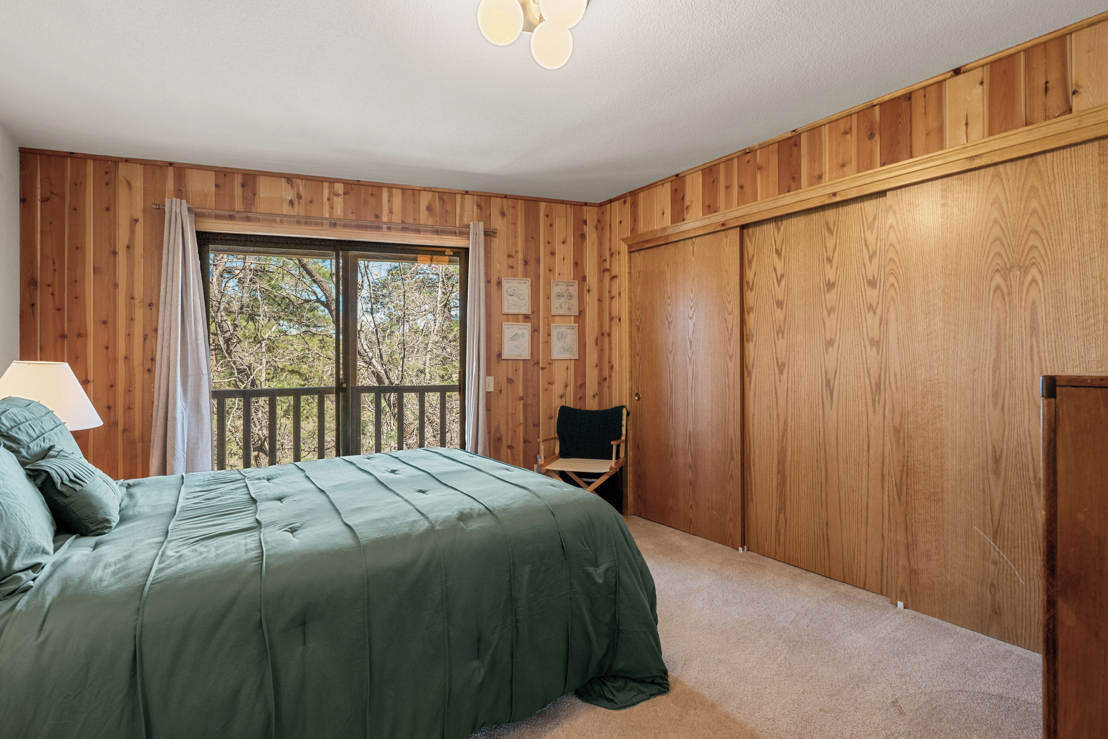Una habitación con paneles de madera, un ventanal grande, una cama con ropa de cama verde, una silla y una lámpara.
