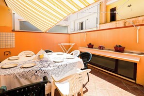 Zona pranzo con tavolo imbandito per quattro persone, sedie, angolo cottura con fornello e finestra vista strada.
