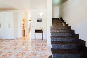 Un corridoio con pavimento piastrellato, una scala, una scrivania con una lampada e un quadro appeso al muro.