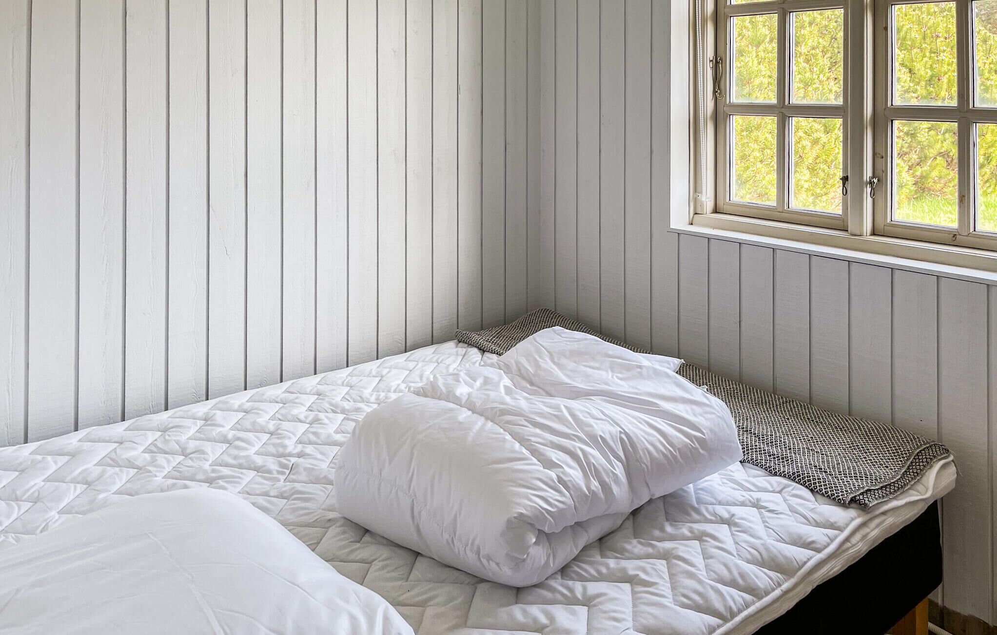 A single bed with white bedding in a room with wooden paneling and a window with a view of greenery.