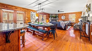 Ein geräumiges Blockhaus-Interieur mit einer Holzeinrichtungs-Essecke, einem Wohnzimmer mit Kamin und einer Küche mit großem Fenster.