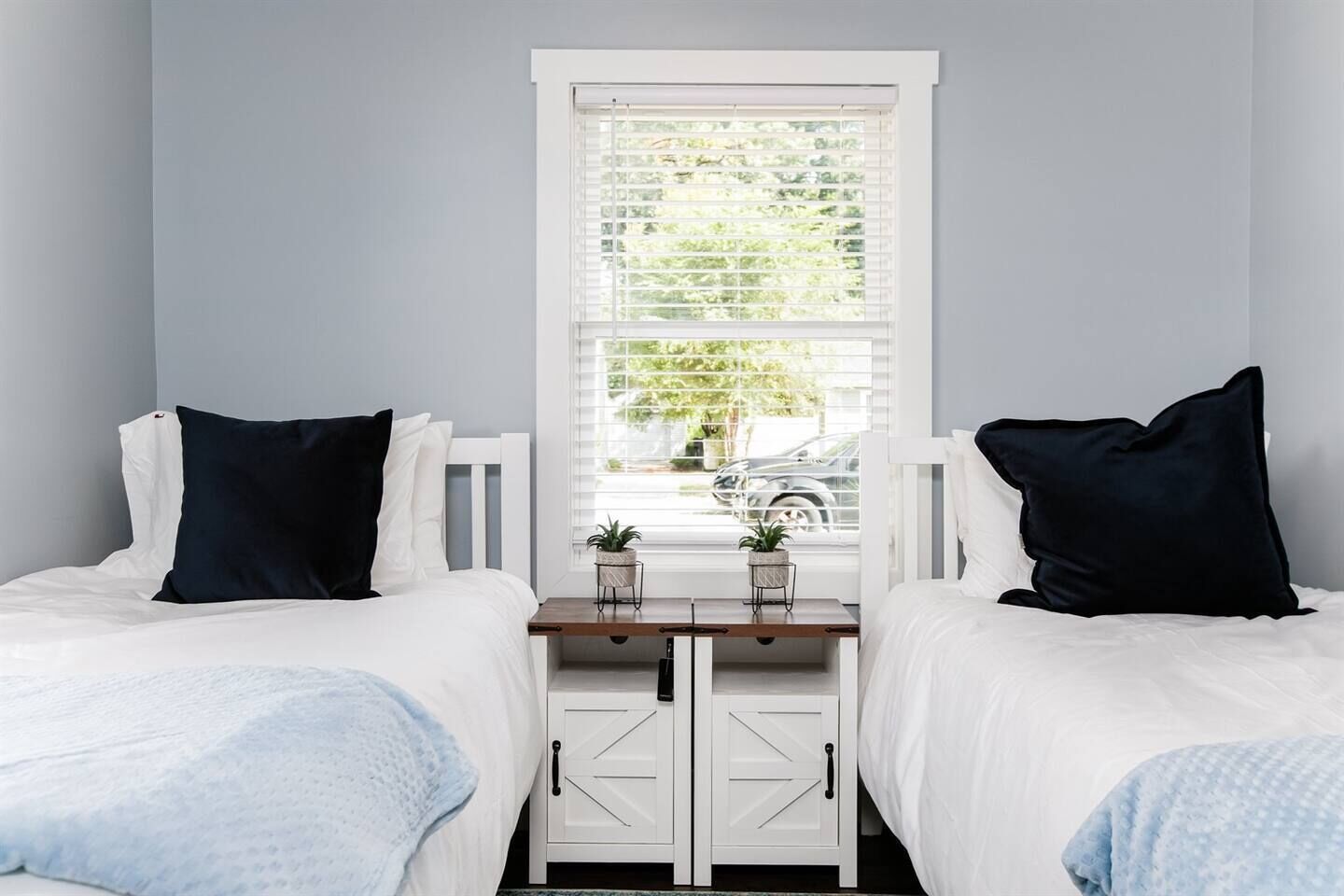 A bedroom with two beds, a window with blinds, a bedside table with a plant, and a wardrobe with a handle.