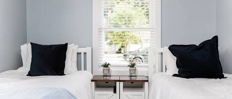 A bedroom with two beds, a window with blinds, a bedside table with a plant, and a wardrobe with a handle.