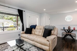 Ein Wohnzimmer mit einem beigen Sofa, schwarzen und blauen Kissen, einem Glastisch und einem dunklen Holzboden.