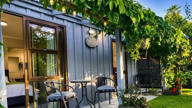Un espace extérieur moderne aménagé avec une porte-fenêtre donnant accès à l’intérieur, une petite table, deux chaises et un jardin avec de la pelouse et des plantes.
