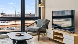 A modern living room with a large window, a flat-screen TV, a grey armchair, a small round table, and a wooden TV stand. - The Edison Old City | Premier 1 BR Suite w/ City View + Shared Roofdeck (Philadelphia)