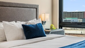 A hotel room with a bed, a nightstand, a lamp, and a window with a view of buildings. - The Edison Old City | Premier 1 BR Suite w/ City View + Shared Roofdeck (Philadelphia)