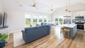 A modern kitchen with a large island, a dining area with a blue sofa, and a TV in the living room. - New Corolla Home with Pool & Spa! (Corolla)