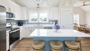 A modern kitchen with white cabinets, stainless steel appliances, a large island with stools, and a view to the dining area. - New Corolla Home with Pool & Spa! (Corolla)