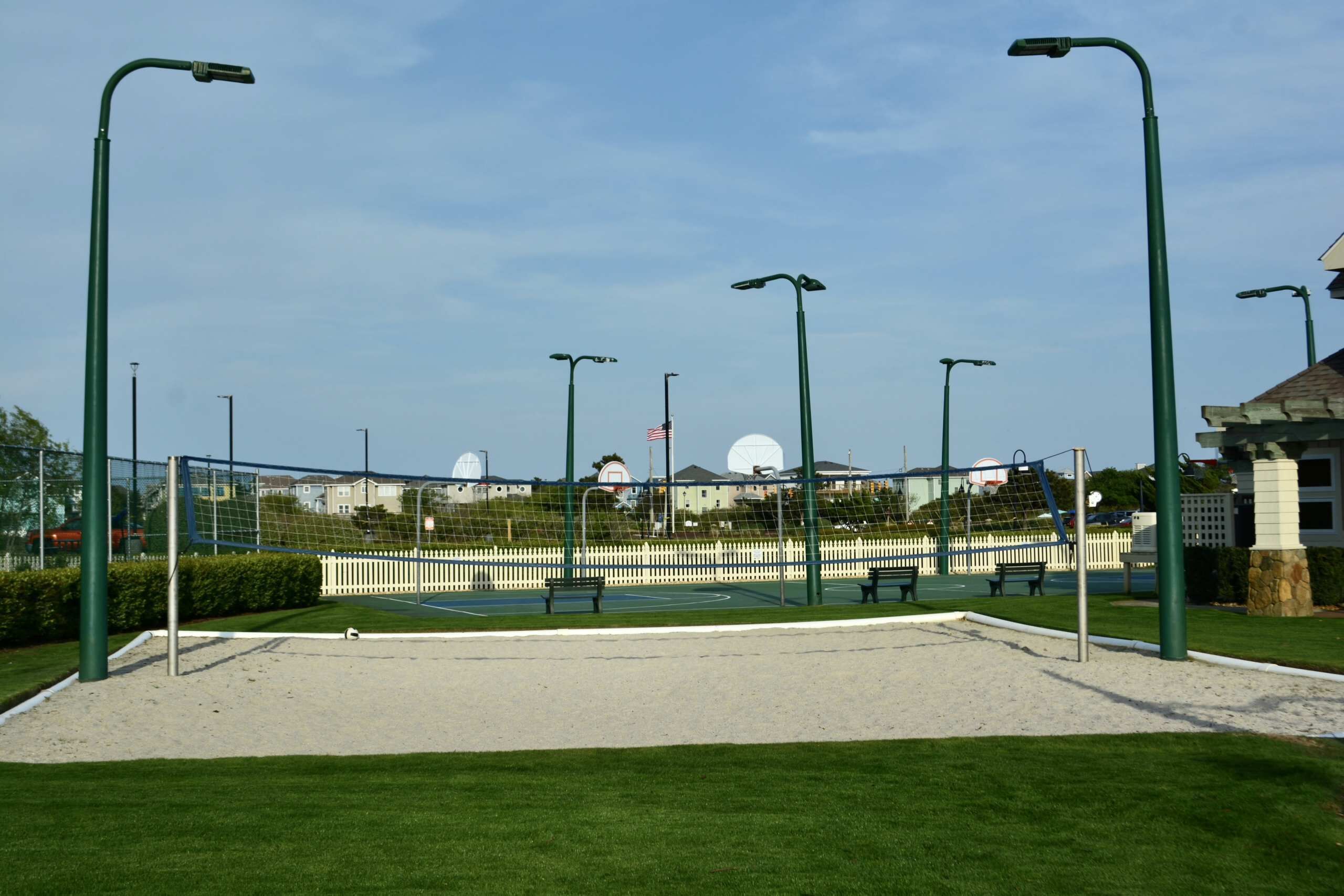 Un court de tennis avec un filet, entouré d’herbe verte et de sable, avec des lampadaires et une clôture en arrière-plan.