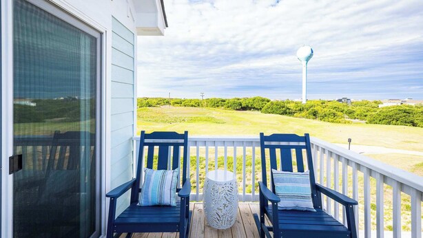A balcony with two blue chairs and a white table, overlooking a grassy area and a water tower. - New Corolla Home with Pool & Spa! (Corolla)