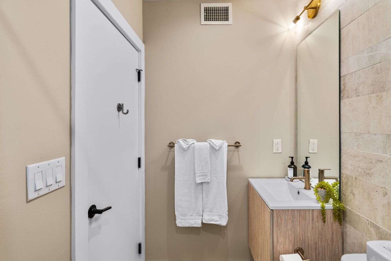 Une salle de bain avec une porte blanche, un porte-serviettes avec deux serviettes, un lavabo avec un miroir et un luminaire au-dessus du miroir.