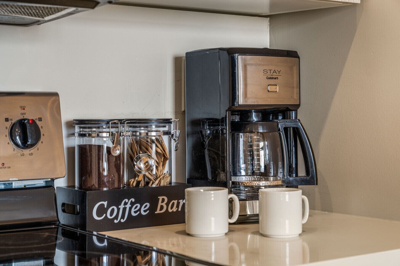 A coffee station with a coffee maker, two coffee mugs, and a container with coffee beans.