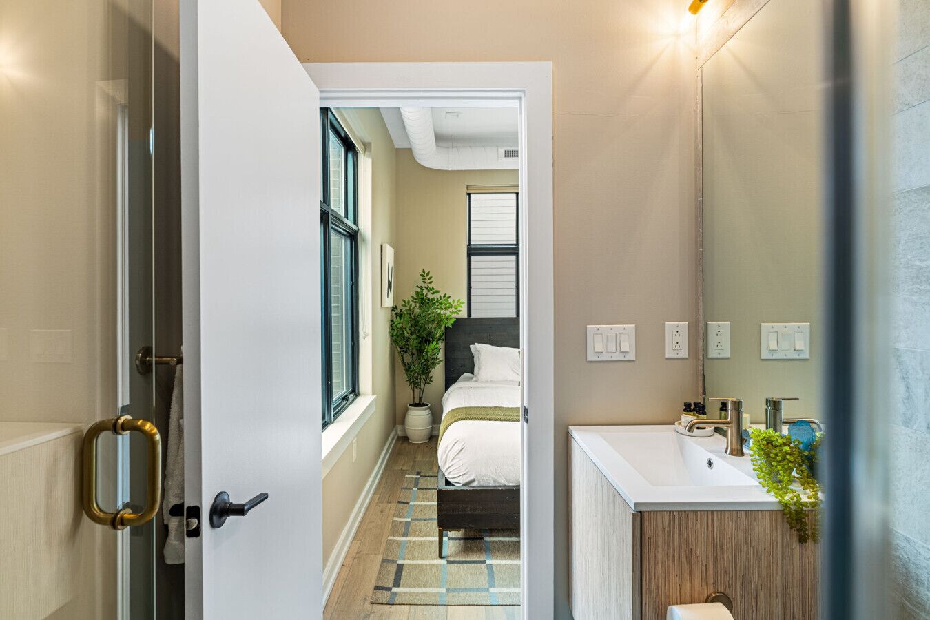 A narrow hallway leading to a bedroom with a bed, a potted plant, and a bathroom with a sink and mirror.