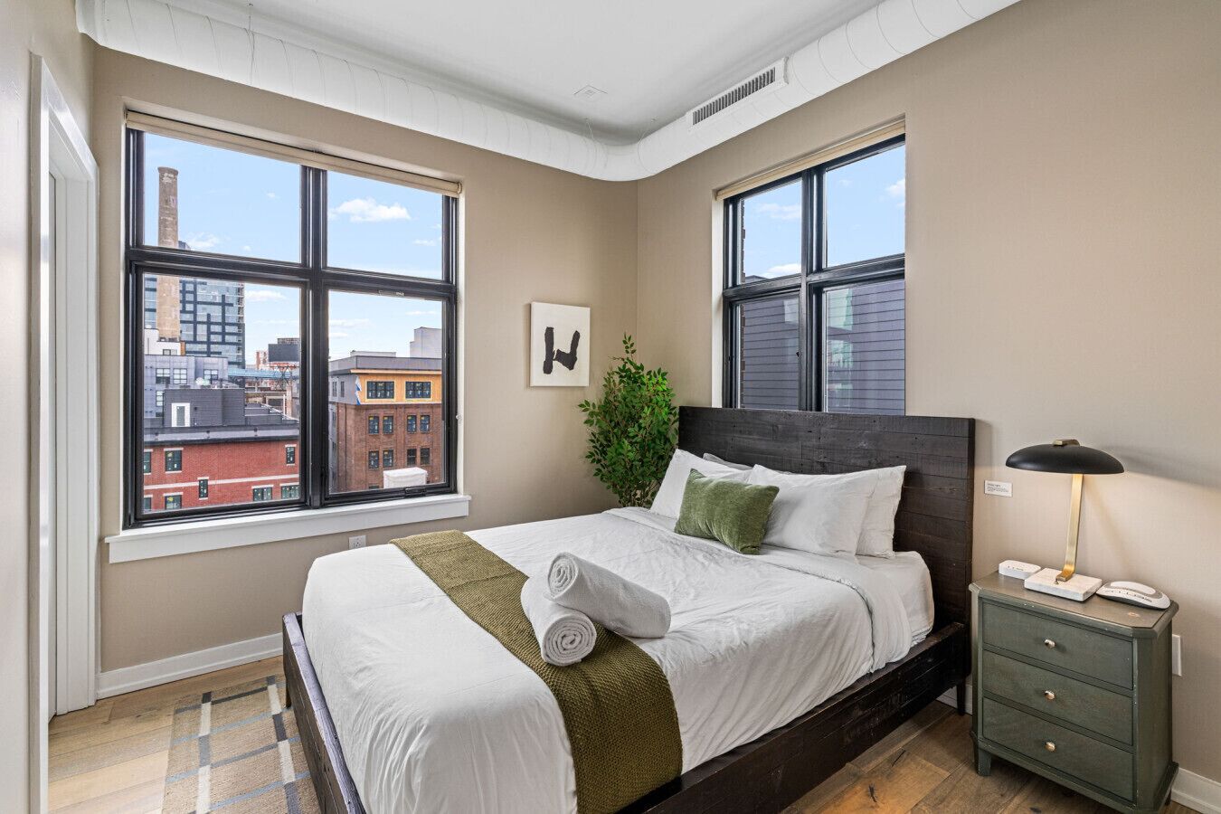 Ein Schlafzimmer mit einem großen Bett, einem Nachttisch, einer Lampe, einer Pflanze und Blick auf Gebäude durch große Fenster.