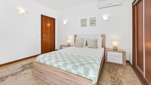 Une chambre à coucher avec un lit en bois, deux tables de chevet avec des lampes, un climatiseur fixé au mur et deux tableaux encadrés accrochés au mur.