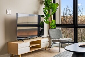 A modern living room with a flat-screen TV on a wooden stand, a grey upholstered chair, and a large green plant. - The Edison Old City | Signature 1 BR Suite w/ Shared Roofdeck (Philadelphia)