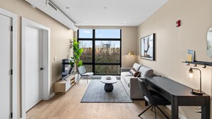 Een moderne woonkamer met een bank, salontafel en televisie. Er is een groot raam met uitzicht op bomen en gebouwen.