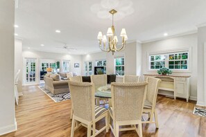 A spacious living area with a dining table surrounded by wicker chairs, a chandelier, and a view of the outdoors through large windows. - Golf Course Corner Villa First Floor Views (Hilton Head Island)