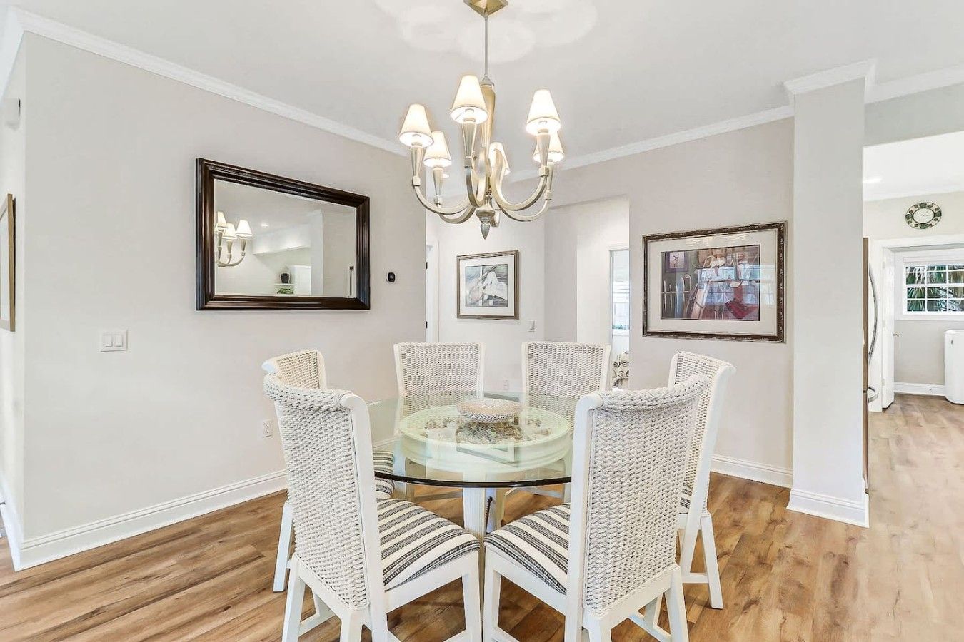 A dining room with a round glass table surrounded by white wicker chairs, a chandelier, and framed artwork on the walls.