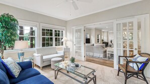 A spacious living room with a sofa, armchair, and coffee table, large windows, and a ceiling fan. - Golf Course Corner Villa First Floor Views (Hilton Head Island)