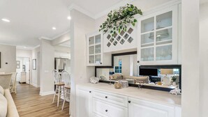 A modern kitchen with white cabinetry, a central island, and a dining area with wicker chairs. - Golf Course Corner Villa First Floor Views (Hilton Head Island)