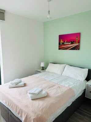 Ein ordentlich bezogenes Bett mit weißen Leinentüchern und einer rosafarbenen Decke, eine Nachttischlampe und ein Stadtbild an der Wand.
