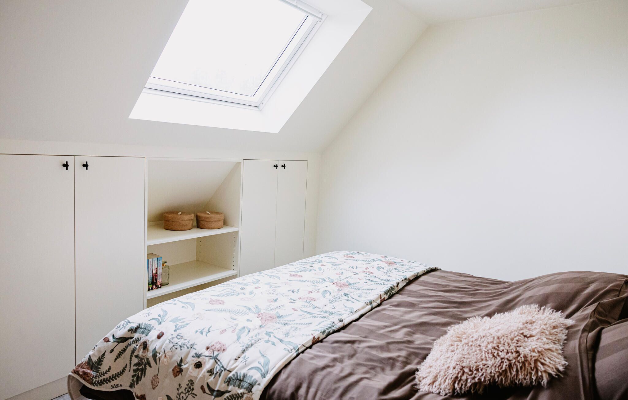 A bedroom with a skylight, a bed with a floral bedspread, a brown headboard, and a white wardrobe with two baskets on the top shelf.