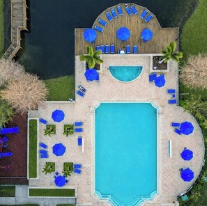 Eine Vogelperspektive auf ein Resort mit einem großen zentralen Pool, zahlreichen Liegestühlen und einer Holzterrasse mit Sonnenschirmen.